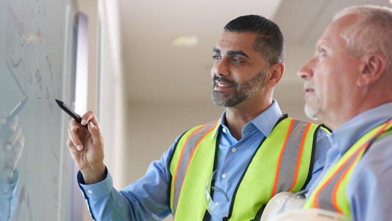 two men in yellow/orange safety vests discussing information whiteboard